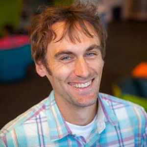 Potrait picture of Jeffrey Bigham, a man with medium-length brown hair, wearing a light shirt with blue and orange checkered stripes, looking at the camera and smiling