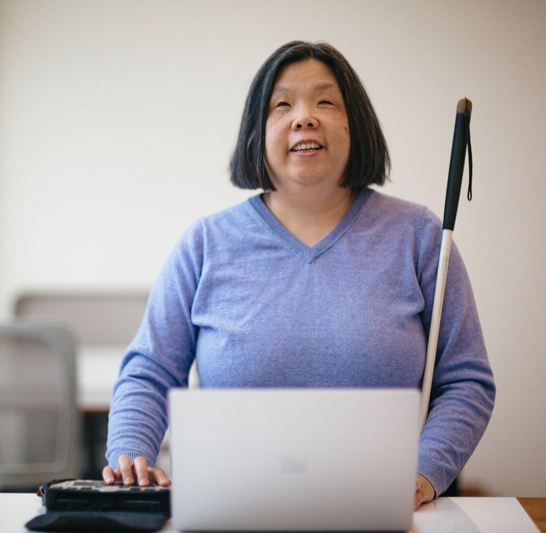 A photo of Anne, a woman with straight black hair, wearing a blue shirt, standing in a classroom, looking directly at the camera smiling