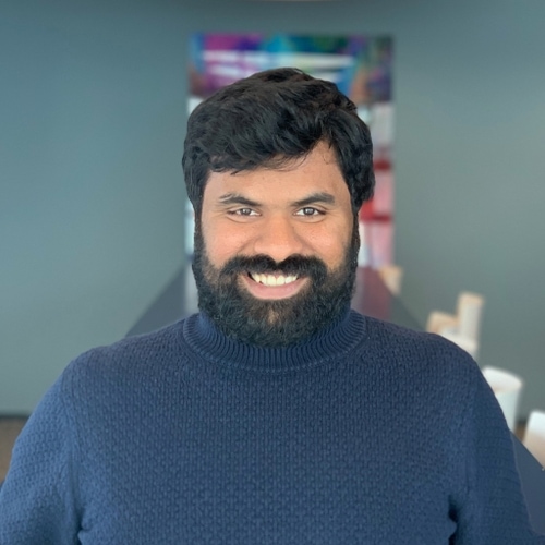A headshot of Karthik, a man with short brown hair and a beard, wearing a dark blue sweater, looking directly at the camera smiling