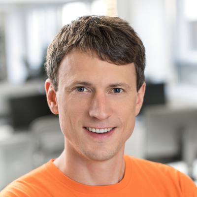 A headshot of Marcus, a man with short brown hair, wearing a bright orange shirt, looking directly at the camera smiling