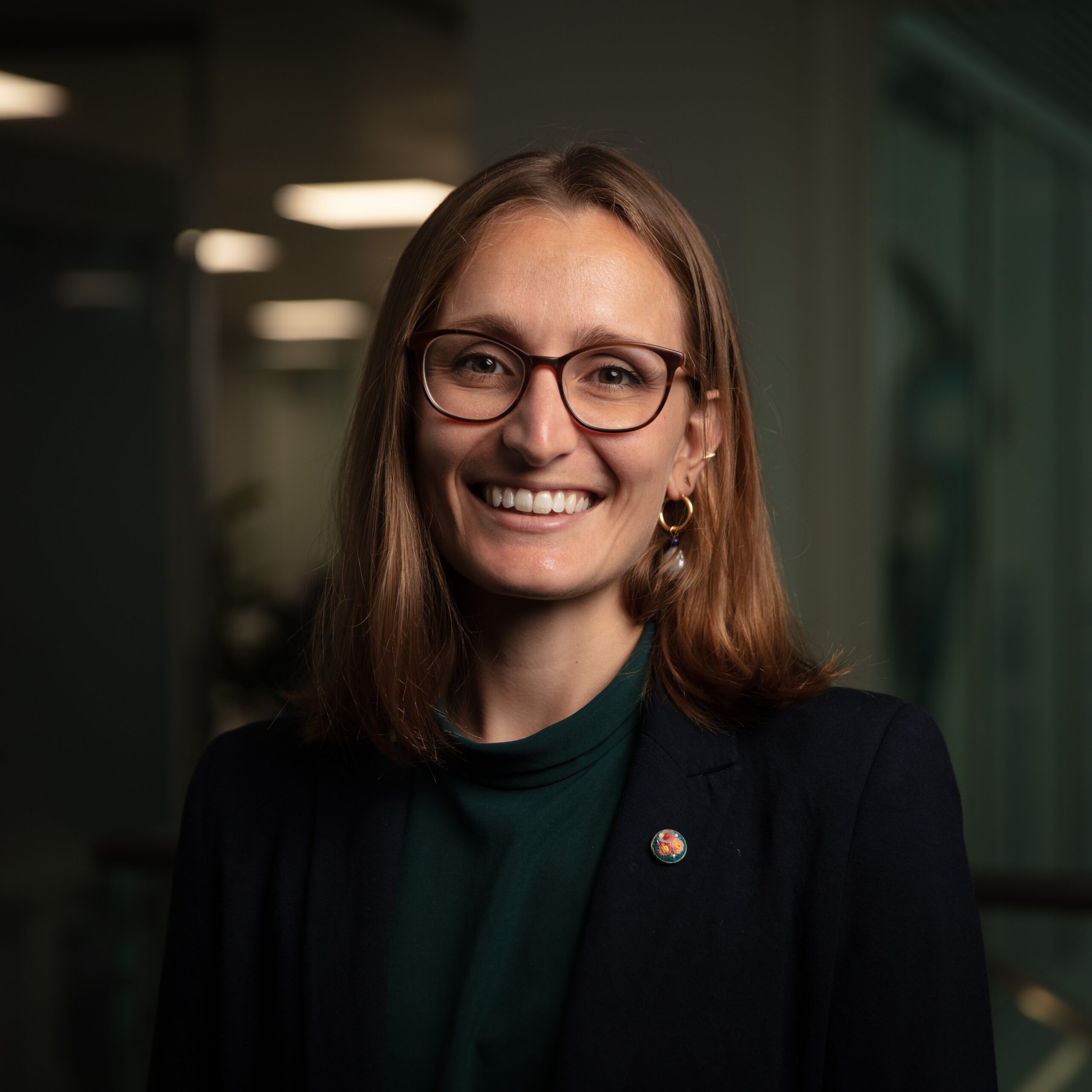 A portrait of Daniela Massiceti, a woman with long auburn hair and a dark business suit. She is wearing glasses and is looking straight at the camera smiling.