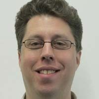 A headshot of Andrew Howard, a man with short dark hair who is wearing glasses, a light shirt and is smiling looking straight at the camera