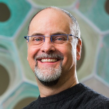 A photo of Ed Cutrell, a man with closely-cropped gray hair and a short beard with mustache. Ed is wearing a black turtleneck and rimless glasses and is looking at the camera smiling.