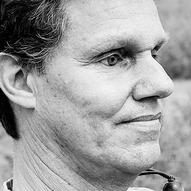 A black and white headshot showing the profile of Daniel, a white man with dark hair, sitting outside and looking at the camera smiling