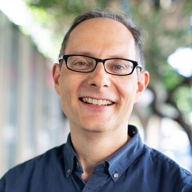 A headshot of James, a man with short brown hair wearing glasses and a dark shirt, standing outside, looking directly at the camera smiling