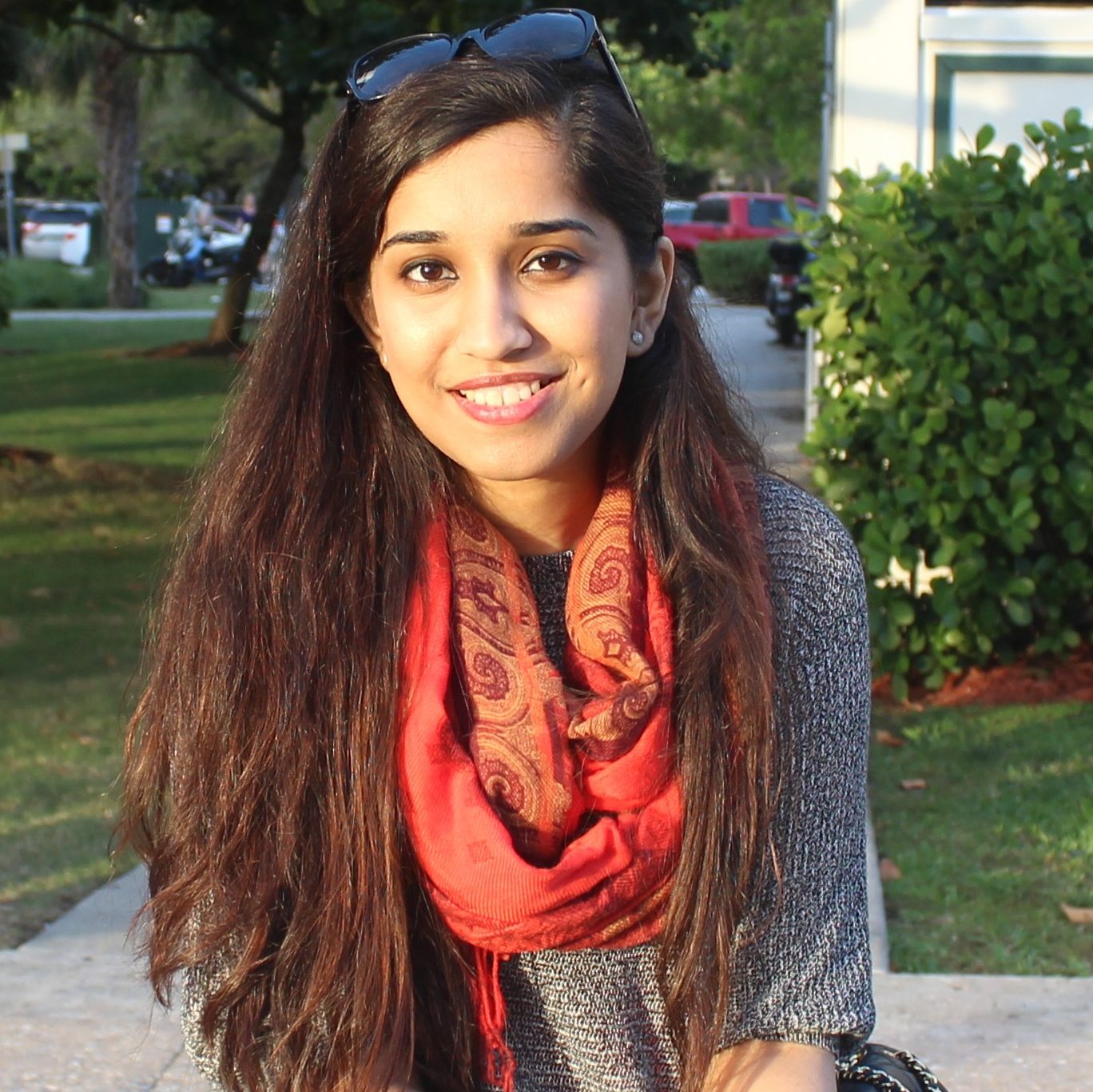 A portrait of Samreen Anjum, a woman with long brown hair, wearing a gray sweater and a red scarf. She is looking at the camera and smiling.