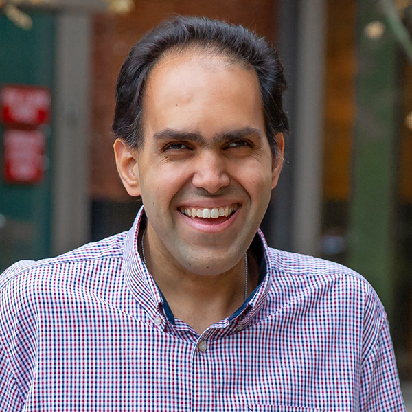 A headshot of Saqib, a man with short dark hair swept back, wearing a checkered reand and blue button-down shirt, looking to the left of the camera smiling