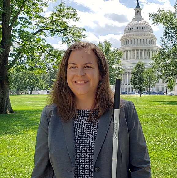 A photo of Stephanie, a woman with long brown hair wearing a grey suit, standing in front of the white house holding a white cane, looking directly at the camera and smiling