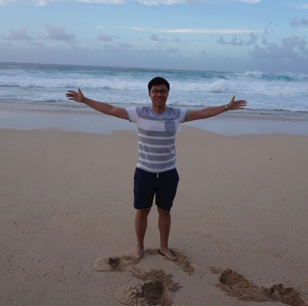 A photo of a man wearing a T-shirt standing on top of a sandy beach in front of the sea.